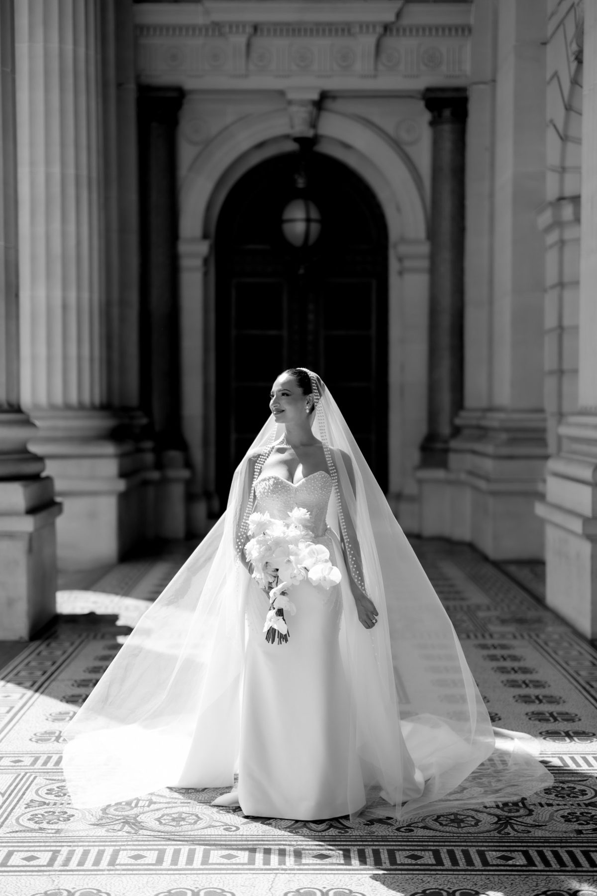Bride standing in a sunlit portico with her veil spread around her