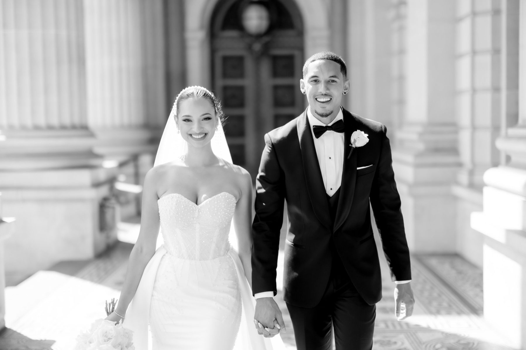 Couple walking toward the camera through a columned portico, both beaming