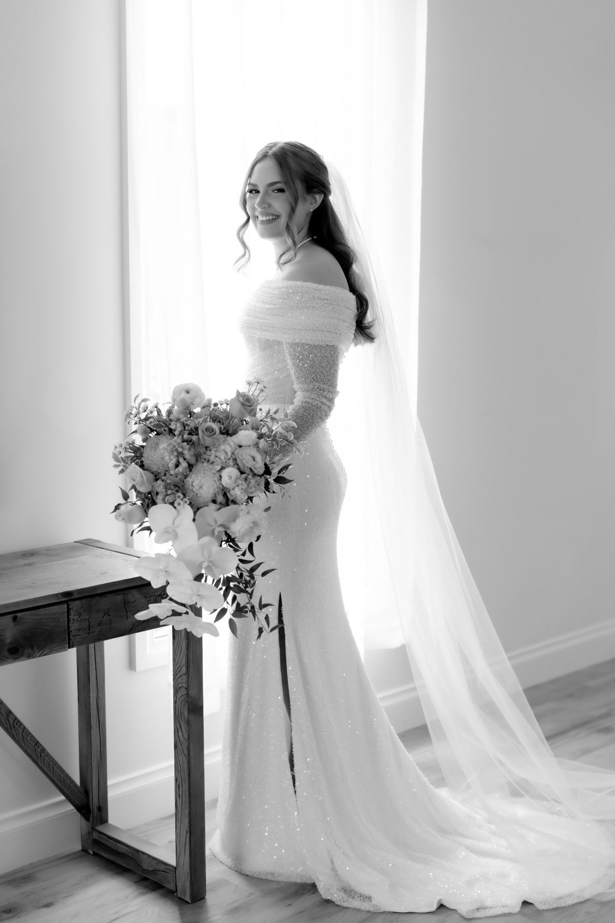 Bride smiling over her shoulder by a bright window, holding her bouquet