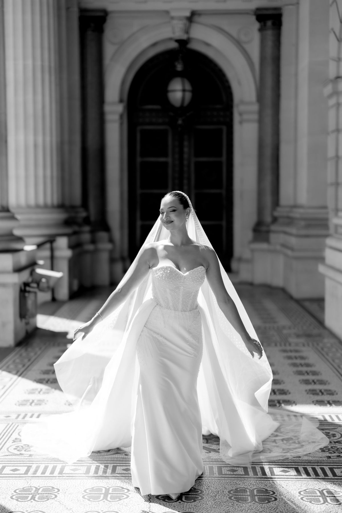 Bride twirling in sunlight beneath a grand stone archway, veil flowing