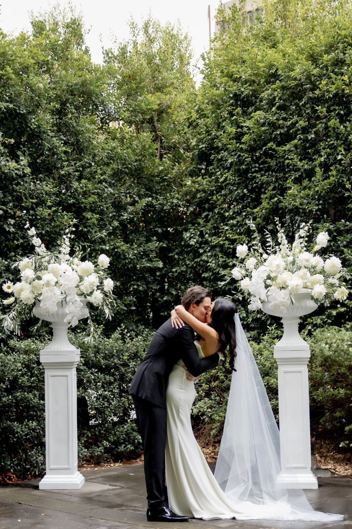 First kiss between two white urns of flowers at the ceremony