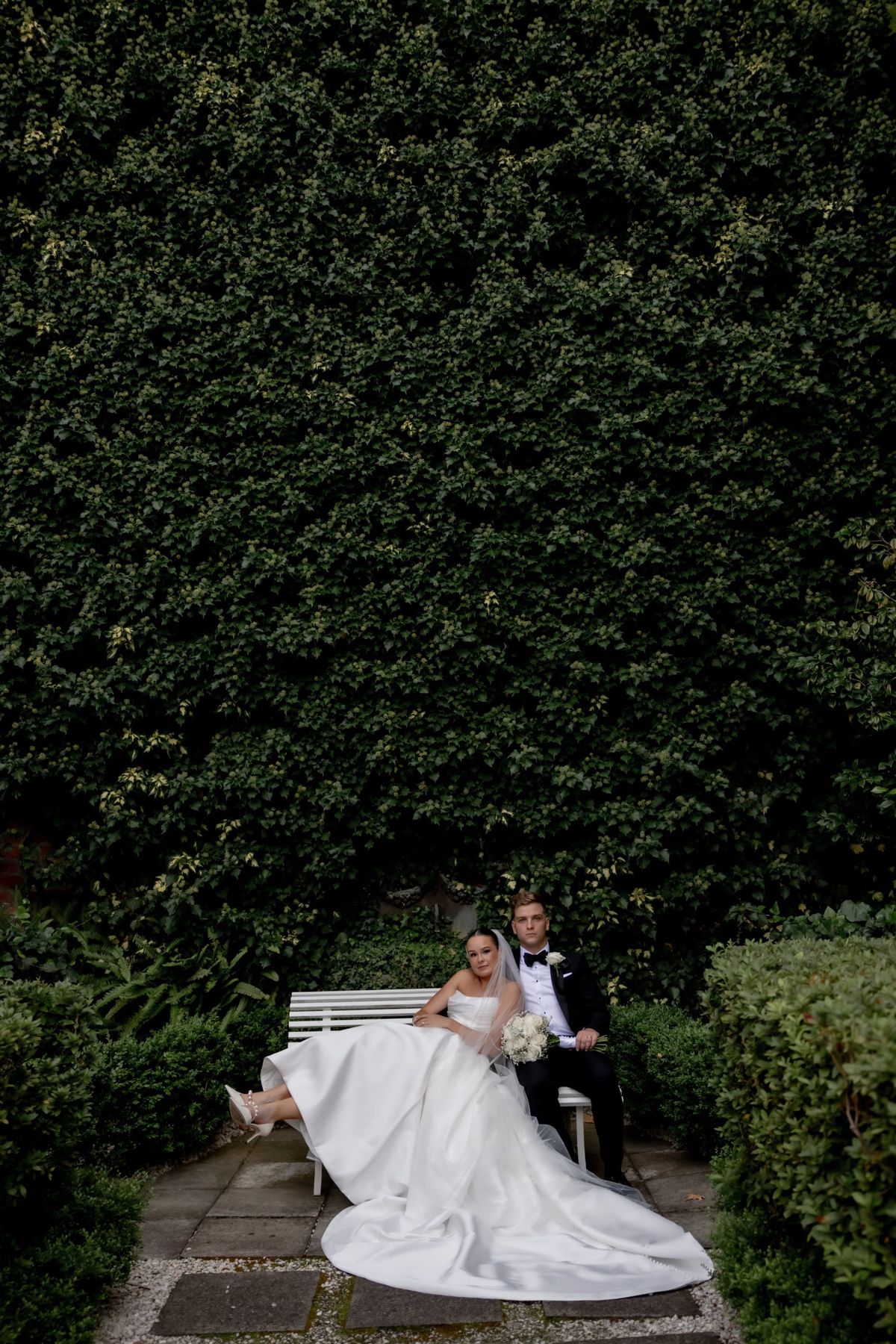 Couple relaxing on a white bench beneath a towering ivy wall