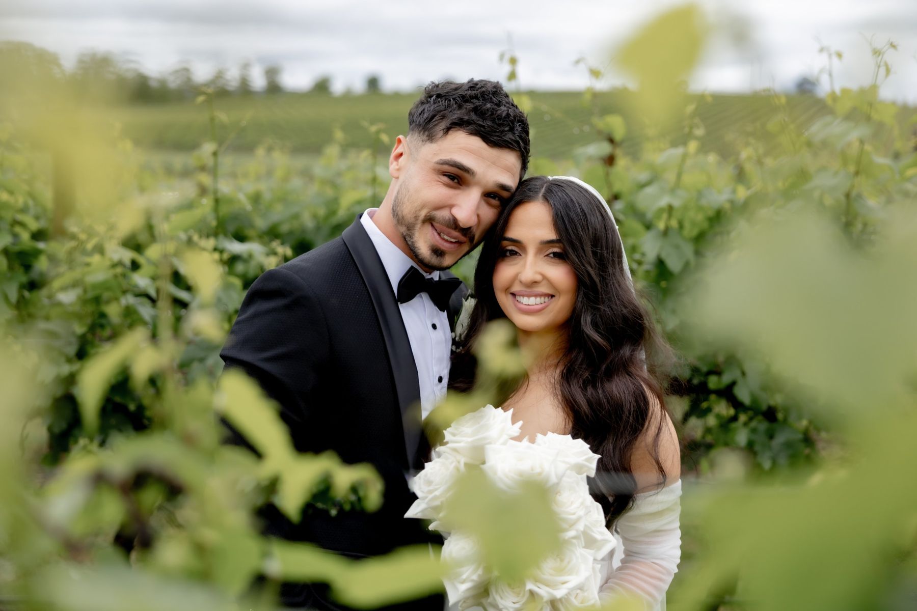 Vineyard wedding photography: couple cheek to cheek, framed by soft vine leaves